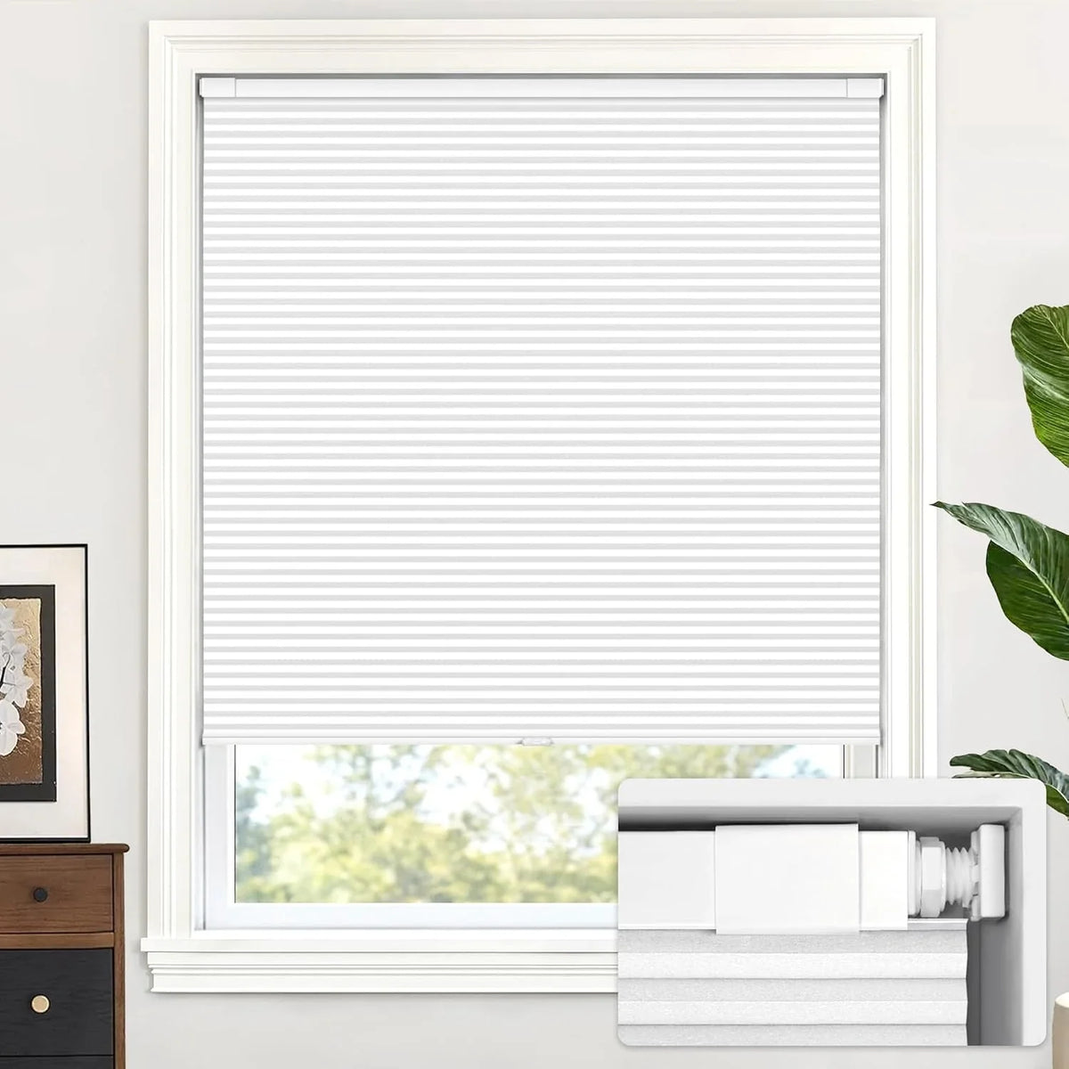 White pleated window blind installed in a white window frame, shown partially drawn with a window view visible below. A product inset at the bottom right shows the blind folded and packaged, and the scene is set in a bright room with a plant and small furniture piece.